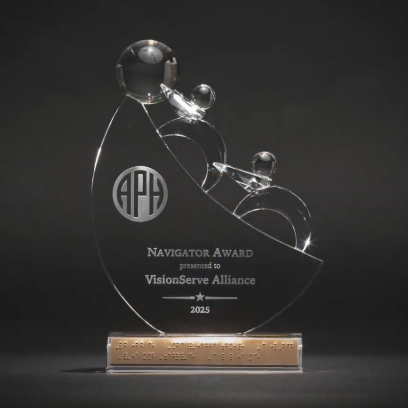 Pictured is the APH Navigator Award from 2025, presented to VisionServeAlliance. The award is a clear glass trophy with a curved, teardrop-like shape with a broad base. Three stylized human figures, each represented by a clear glass sphere for a head and curved geometric shapes for the body, ascend upward together along the curved shape of the award. Near the center of the trophy is a circular APH logo.