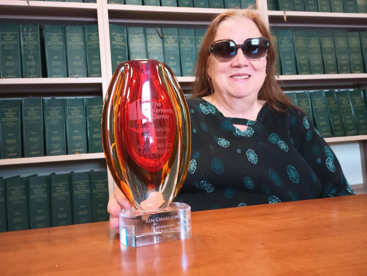 Kim sits in a library surrounded by braille books as she proudly smiles while holding a red crystal award.