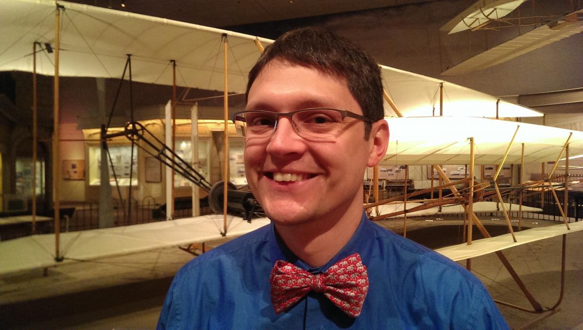 A light-skinned man with half-rim glasses and short brown hair wears a bright blue shirt with contrasting red bowtie and stands in front of the 1903 Wright Flyer while smiling broadly at the camera.