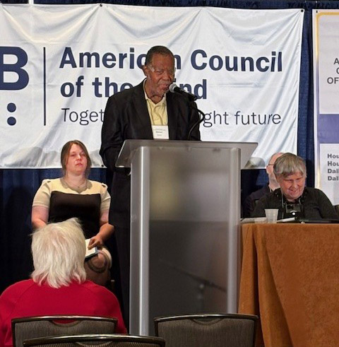 Treasurer Michael Garrett behind a clear podium. Right behind him is a white banner in dark blue lettering is the American Council of the Blind logo and slogan "Together for a bright future". Seated on the back left of Michael is one of the Texas thrift store managers. On his back right is the Thrift Store General Store manager. Seated on Michael's lower right is President Deb Cook Lewis behind a clothed table. A member in red is seated on a table on in front of the thrift store manager on a lower level.