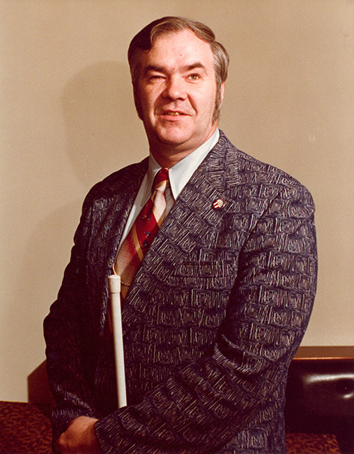 Oral Miller stands tall, sporting a dark suit, white shirt, and red-and-white tie, with a USABA pin on his left lapel.